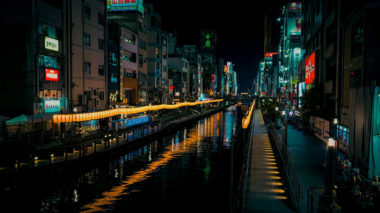 A canal in Osaka at night: a long string of yellow lanterns along one bank, neon signs above, their light doubled in the black water.