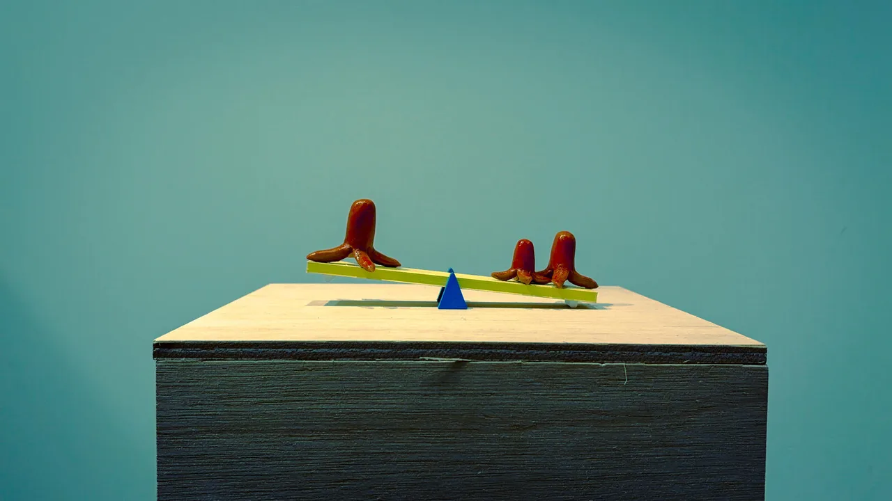 Three red octopus-shaped sausages balanced on a tiny yellow see-saw, on a plywood plinth against a teal wall.