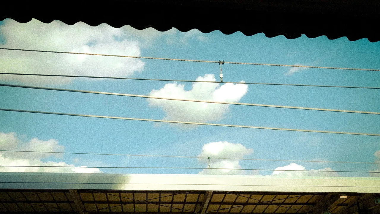 A strip of blue sky and white clouds between a scalloped awning and the edge of a roof, crossed by power lines.