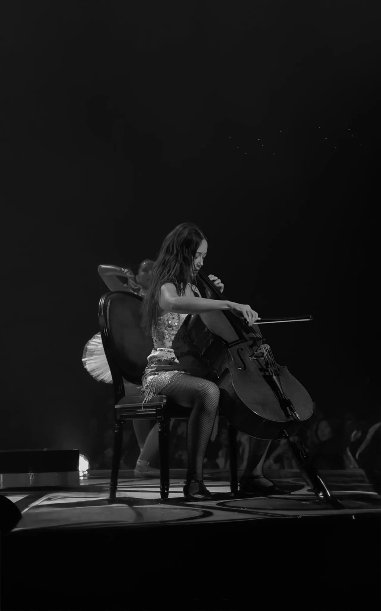 Black and white, upright: Laufey on a dark stage, playing the cello and singing into a microphone.