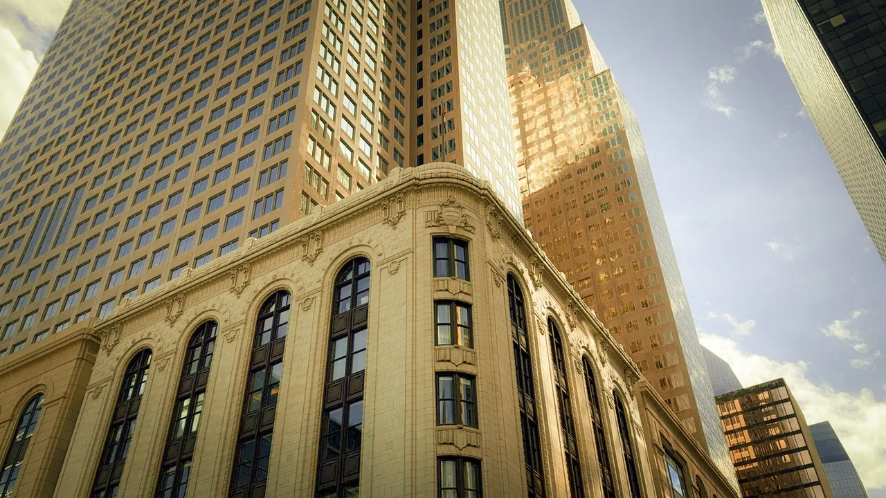 Looking up at a cream stone building with arched windows, set against golden glass towers and a bright, cloudy sky.