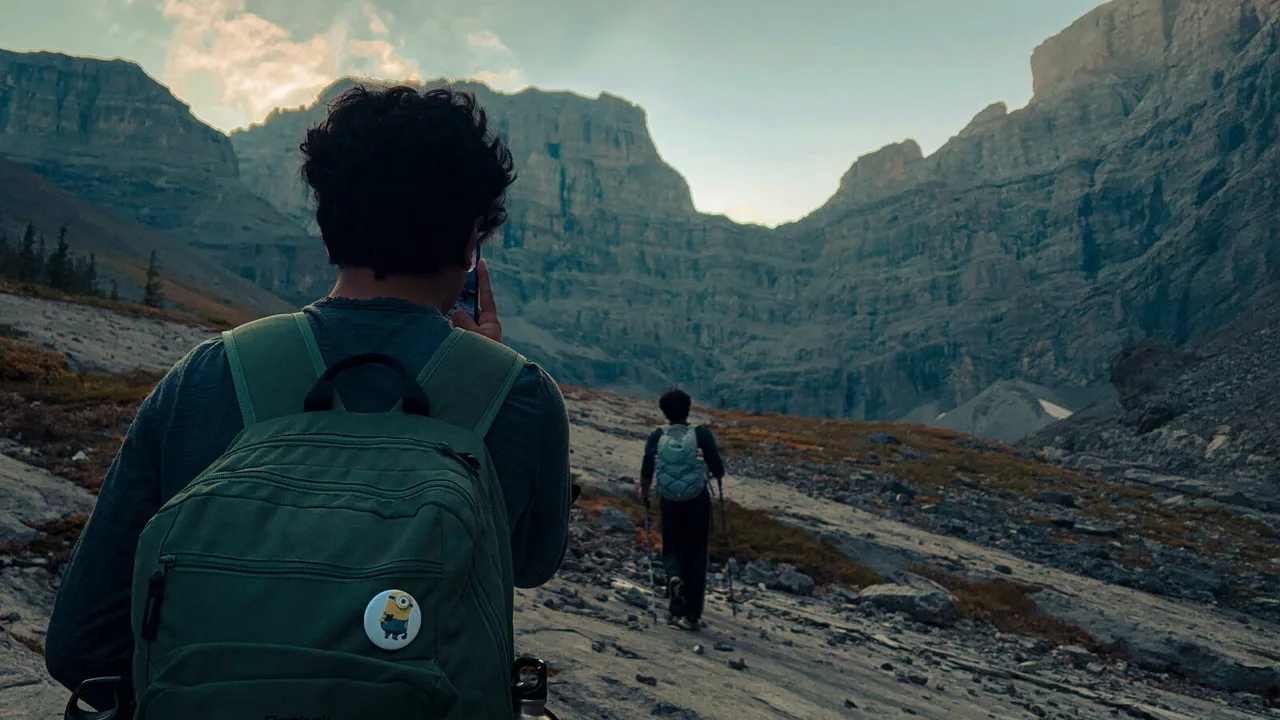 A hiker with a green backpack photographs a wide, rocky valley ringed by cliffs, while another walks on ahead.