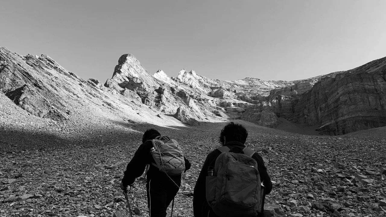 Black and white: two hikers with packs climb a slope of loose stones toward jagged peaks.