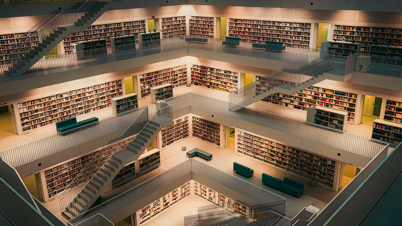 Looking down into a white library atrium, tier upon tier of bookshelves linked by staircases, a few teal sofas far below.